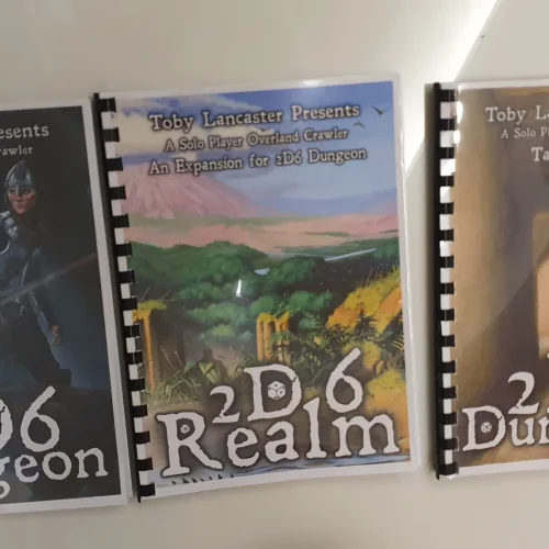 Set of comb bound gaming rulebooks with illustrated covers — printed guides for the 2D6 Dungeon tabletop game series using black plastic comb binding.
